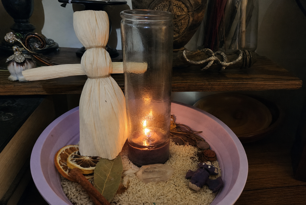 Mabon prosperity altar with a corn husk doll, candle, rice, dried orange, cinnamon, bay leaves, coins, and herbs.